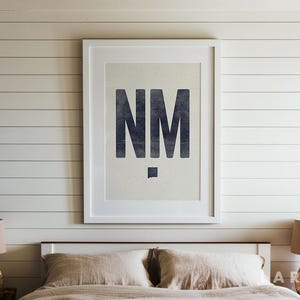 May include: A framed print featuring the letters "NM" in a dark blue, distressed font. The artwork is centered on a light beige background, set within a white frame. The print hangs on a white shiplap wall above a bed with neutral-toned bedding.