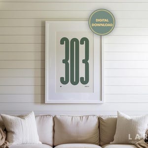 May include: A framed art print featuring the number "303" in a bold, dark green font. The artwork is set against a white background within a white frame. A "DIGITAL DOWNLOAD" sticker is visible. The print hangs above a beige sofa with white pillows.