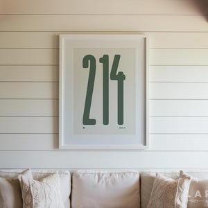 May include: A framed print with the number "214" in a dark green font. The print is centered on a white wall above a white couch with decorative pillows. The room has a modern, minimalist aesthetic.
