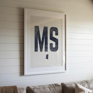 Op de afbeelding: Een ingelijste print met de letters "MS" in een donkerblauwe, vette letter. De print staat tegen een lichte achtergrond en is omlijst met een witte rand. Het kunstwerk is gemonteerd op een witte lambrisering.