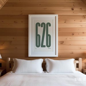 May include: A bedroom interior with a bed dressed in white bedding and pillows. A framed print displaying the number "626" in green is mounted above the bed. Two bedside lamps with white shades are placed on wooden bedside tables.