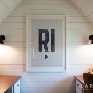 Può includere: Una stampa incorniciata con le lettere "RI" in blu scuro, con un contorno della mappa del Rhode Island sottostante. La stampa è appesa a una parete bianca con doghe, affiancata da due applique nere. Una scrivania in legno è visibile sotto.
