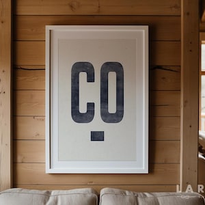 Puede incluir: Un estampado enmarcado con las letras "CO" en azul oscuro, con un estilo desgastado. La obra de arte est&aacute; sobre un fondo beige claro y enmarcada en un borde blanco. El estampado est&aacute; montado en una pared de madera.