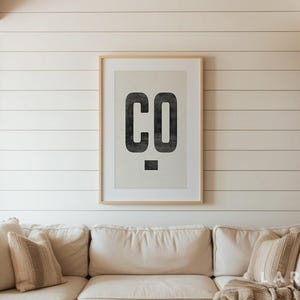 Puede incluir: Un estampado enmarcado con las letras "CO" en una fuente negra audaz y desgastada. La obra de arte se exhibe en una pared blanca de tablillas sobre un sof&aacute; color crema con cojines decorativos. La est&eacute;tica general es moderna y r&uacute;stica.