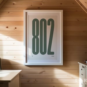May include: A framed print with the number "802" in a bold, green font against a white background. The artwork is displayed on a light wood-paneled wall, creating a minimalist aesthetic. The print is in a white frame.
