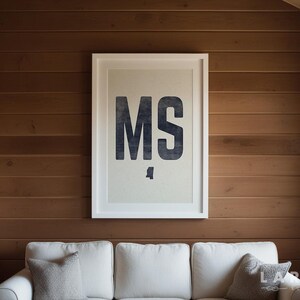 Op de afbeelding: Een ingelijste print met de letters "MS" in donkerblauw, met een silhouet van de staat Mississippi eronder. De print, in een witte lijst, hangt aan een houten wand boven een witte bank met grijze kussens.