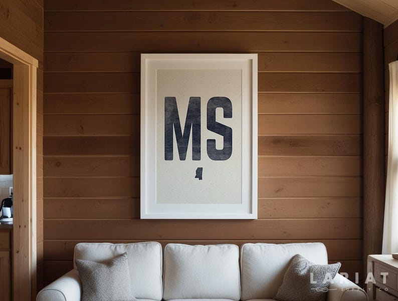 Op de afbeelding: Een ingelijste print met de letters "MS" in donkerblauw, met een silhouet van de staat Mississippi eronder. De print, in een witte lijst, hangt aan een houten wand boven een witte bank met grijze kussens.