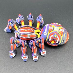 May include: A multi-colored mechanical spider figurine featuring a purple, red, and yellow color scheme with articulated legs and a separate, rounded, intricately detailed shell component positioned next to the main body of the spider figure.