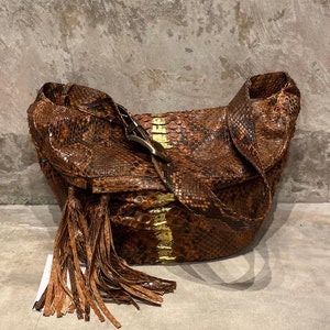 Pode incluir: Uma bolsa de mão de pele de cobra marrom com um detalhe de franja e uma fivela dourada. A bolsa está sobre uma superfície de concreto.
