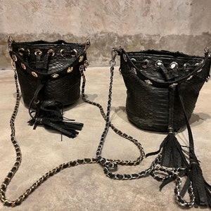 Könnte beinhalten: Zwei schwarze Ledereimer-Taschen mit goldenen Kettenriemen und schwarzen Quasten. Die Taschen haben ein Schlangenhautmuster und goldene Nieten am oberen Rand.