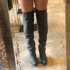 Puede incluir: Un par de botas negras de caña alta con estampado de piel de serpiente. Las botas tienen punta puntiaguda y tacón de bloque.