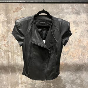 May include: Black leather jacket with short sleeves and a zip closure. The jacket has a collar and a label that says "REMARK" on the inside.