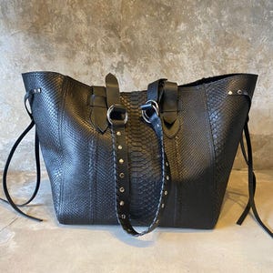 Könnte beinhalten: Eine schwarze Ledertasche mit Schlangenhautmuster und silbernen Beschlägen. Die Tasche hat zwei obere Henkel und einen langen, verstellbaren Riemen.