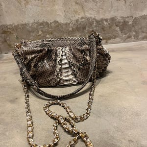 Könnte beinhalten: Eine kleine, graue und weiße Schlangenleder-Handtasche mit einer goldenen Kettenriemenschlaufe.