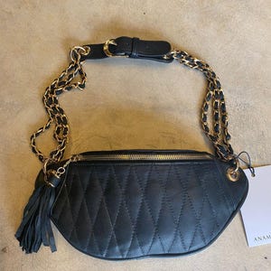 Könnte beinhalten: Schwarze gesteppte Bauchtasche mit goldener Kettenkette und Quaste. Die Tasche hat einen Reißverschluss und eine goldene Schnalle.
