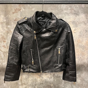 Könnte beinhalten: Schwarze Lederjacke mit Schlangenhautmuster und goldfarbenen Reißverschlussdetails. Die Jacke hat einen Reißverschluss, zwei Reißverschlusstaschen und einen Gürtel an der Taille.