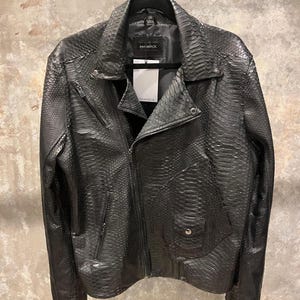 Puede incluir: Una chaqueta de cuero negra con estampado de piel de serpiente. La chaqueta tiene cierre de cremallera y cuello. El nombre de la marca "ANNAMARC" es visible en la etiqueta.