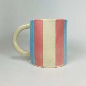 May include: A ceramic mug with a rounded handle. The mug features vertical stripes in light blue, pink, and off-white. The mug is set against a plain white background.