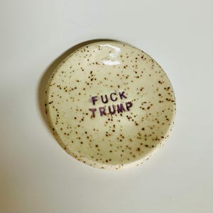 May include: A speckled, off-white ceramic dish with the phrase "FUCK TRUMP" in purple lettering. The dish is round with a slightly raised edge.