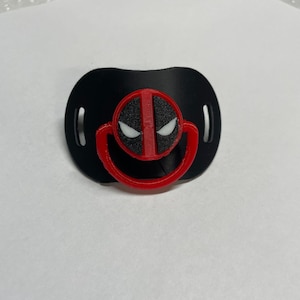 Könnte beinhalten: Schwarz-roter Babyschnuller mit dem Deadpool-Logo. Der Schnuller hat eine schwarze Basis mit roter Kontur und ein rotes Deadpool-Maskendesign in der Mitte. Der Schnuller ist für Säuglinge konzipiert.