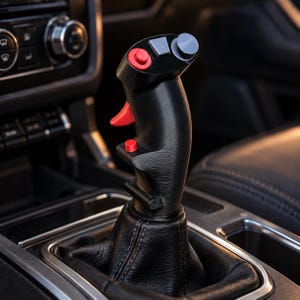 Puede incluir: Un pomo de palanca de cambios negro con forma de palanca de control de vuelo, que presenta un gatillo rojo, un botón rojo en el lateral y un panel superior con un botón rojo, un botón rectangular gris y un joystick circular gris.