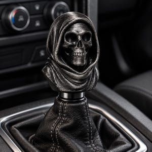 May include: A gear shift knob featuring a detailed, dark grey, weathered skull wearing a hooded cloak. The skull is mounted atop a black leather shift boot with visible white stitching, positioned within a vehicle's center console interior.