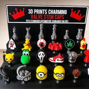 Puede incluir: Un expositor negro escalonado con varios tapones de válvula impresos en 3D para válvulas Schrader. El letrero dice 3D PRINTS CHARMING VALVE STEM CAPS FITS STANDARD AUTOMOTIVE SCHRADER VALVES. Los tapones presentan diversas formas, iconos y figuras.