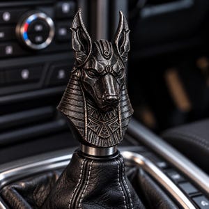 May include: A gear shift knob featuring a detailed, metallic, dark grey sculpture of the Egyptian deity Anubis. The head of the jackal-headed figure is mounted atop a black leather shift boot with white stitching, set within a vehicle interior.