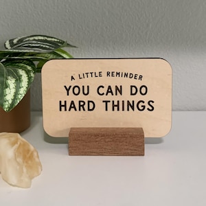 May include: A wooden desk sign with the text "A LITTLE REMINDER YOU CAN DO HARD THINGS" in black lettering. The sign is on a small wooden stand. A potted plant and a decorative rock are in the background.