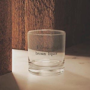 May include: A clear, lowball glass with the etched text "I drink that brown liquor." The glass is cylindrical and sits on a white surface, with a wooden background. The lighting creates shadows, highlighting the glass's shape and the text.