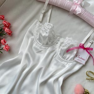 May include: A white satin slip with lace trim, adjustable straps, and a tag that reads "Calméa". The slip is displayed on a pink satin hanger with a bow. A pink makeup brush and gold-colored scissors are also visible.