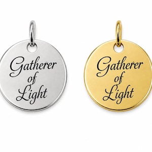 May include: Two circular metal pendants, one silver-toned and one gold-toned, each featuring a jump ring at the top. Both pendants are engraved with the black script text Gatherer of Light centered on the front surface of the smooth metal discs.