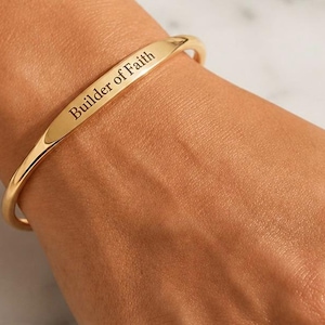 May include: Gold-coloured cuff bracelet inscribed with "Builder of Faith." The curved bracelet is worn on a wrist. The text is in a simple, elegant font.