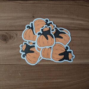 Puede incluir: Una colección de pegatinas de calabazas naranjas con siluetas de gatos negros. Las pegatinas tienen un borde blanco y están dispuestas sobre una superficie de madera. Las calabazas tienen líneas verticales y los gatos están en varias poses.