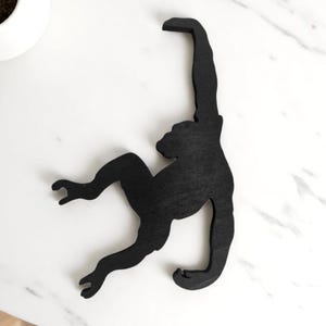 Puede incluir: Silueta de mono de madera negra, colgando de una superficie. El mono está en una pose juguetona, con los brazos y las piernas extendidos. El fondo es una superficie de mármol blanco con una pequeña planta en maceta.