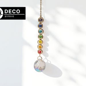 Könnte beinhalten: Ein dekorativer, hängender Kristall-Sonnenfänger mit einer goldenen Kette. Der Sonnenfänger hat eine Reihe von bunten, runden Perlen in einem Regenbogenspektrum, gekrönt von einer klaren, facettierten Kristallkugel. Das Logo "DECO BYPRIVE" befindet sich in der oberen linken Ecke.