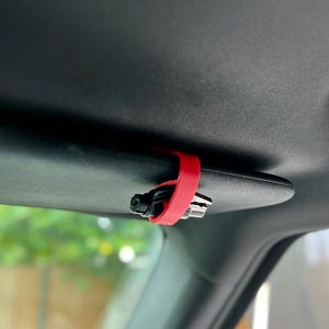 Op de afbeelding: Een felrode plastic clip bevestigd aan de zwarte zonneklep van een voertuig. De clip houdt een set metalen sleutels stevig vast tegen de onderkant van de zonneklep, waardoor ze georganiseerd en toegankelijk blijven in het interieur van de auto.