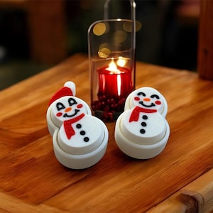 May include: Two white snowman-shaped bottle stoppers with red scarves and black eyes and buttons. The stoppers are on a wooden surface with a candle in a glass jar in the background.