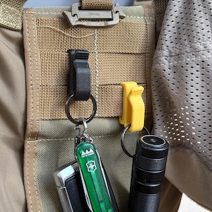 Puede incluir: Una mochila táctica de color tostado con correas MOLLE, que incluye un clip de plástico negro que sujeta una multiherramienta verde y una llave de coche, junto a un clip amarillo unido a una linterna cilíndrica negra sobre la tela exterior.