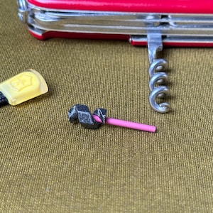 Op de afbeelding: Een rood Zwitsers zakmes met een zilveren kurkentrekker en een zwart koord met een gele sluiting. Er is ook een klein, donkergrijs gereedschap met een roze staaf aanwezig.
