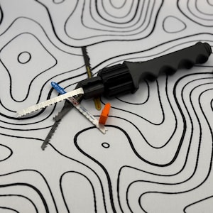 May include: A black coping saw with a gray handle and several saw blades. The blades are silver and have various colors on the ends. An orange blade holder is also included. The items are on a white surface with black contour lines.