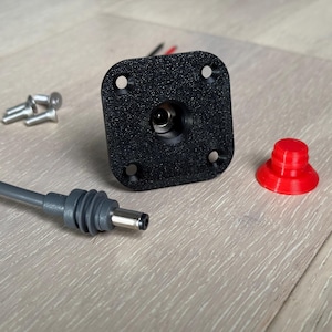 May include: A black plastic electrical outlet with a round hole in the centre. A grey electrical cord with a plug is connected to the outlet. A red plastic cap is also shown.