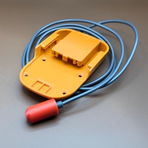 Op de afbeelding: Een gele plastic batterijlader met een blauwe kabel en een rode stekker.