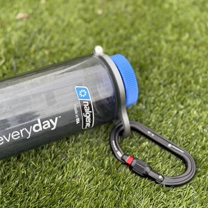 Może przedstawiać: Szara butelka Nalgene z niebieską zakrętką i czarnym karabińczykiem. Na butelce widnieje napis "everyday" i logo Nalgene. Butelka leży na zielonej trawie.