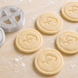 May include: A silver-coloured circular cookie cutter and a matching stamp are positioned next to seven round, light-coloured dough discs. Each dough disc features a debossed design of a stylised cloud with a line extending downward, matching the cutter.