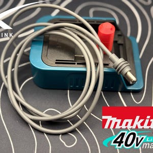 May include: A gray power cord with a red connector is plugged into a teal blue Makita 40V Max XGT Li-ion battery charger.