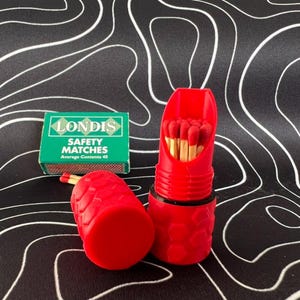 Puede incluir: Dos porta-cerillas rojos con una caja de cerillas de seguridad Londis. Los porta-cerillas están llenos de cerillas con punta roja.