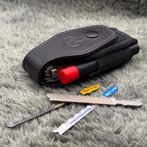 Könnte beinhalten: Ein schwarzes Leder-Multitool-Etui mit einer schwarzen Nylontasche und einem rotspitzigen Werkzeug. Drei Metallsägeblätter und eine Feile mit gelben und blauen Etiketten werden auf einer grau strukturierten Oberfläche gezeigt.
