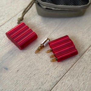Könnte beinhalten: Zwei rote, gerippte Halterungen für kleine Ampullen, mit mehreren sichtbaren Ampullen. Eine kleine, olivgrüne Tasche befindet sich im Hintergrund. Die Artikel sind auf einer hellen Holzoberfläche angeordnet.