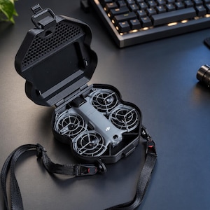 Könnte beinhalten: Eine graue DJI-Drohne mit vier Propellerschutzvorrichtungen liegt in einem schwarzen, geöffneten, muschelförmigen Transportkoffer. Der Koffer hat ein strukturiertes Wabenmuster auf dem Innendeckel und einen schwarzen Trageriemen.